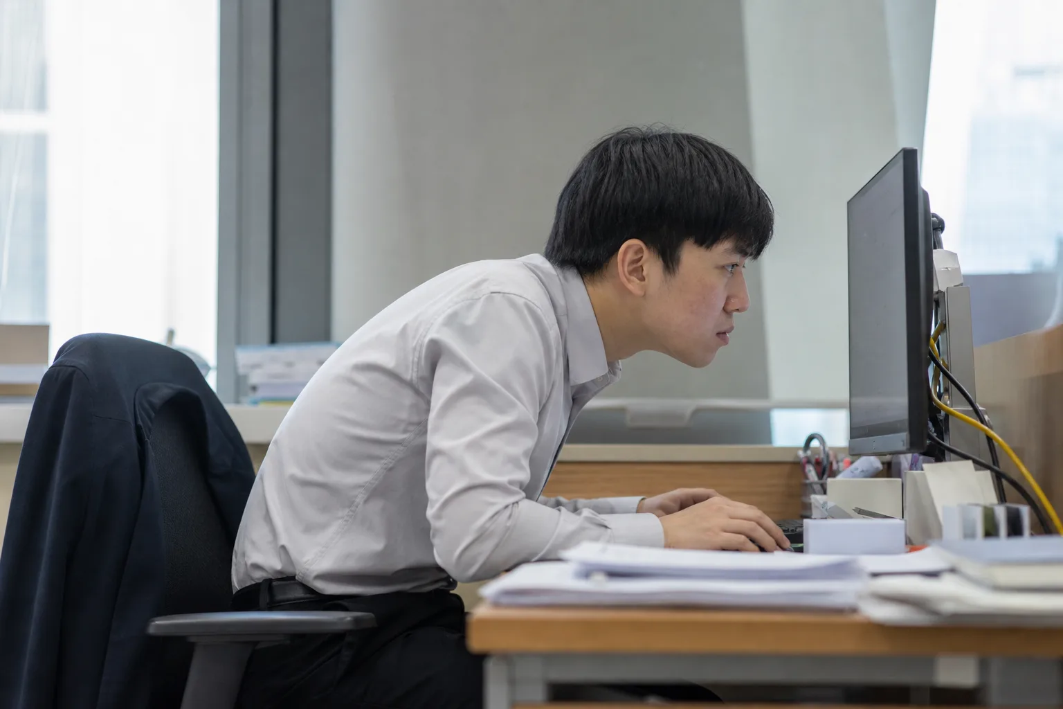 모니터 앞에서 목을 앞으로 빼고 어깨를 웅크린 채 일하는 사무직 직장인 — 승모근 긴장과 반복되는 어깨 담의 원인 자세, 고양 향동 경희미르애한의원 통증 칼럼